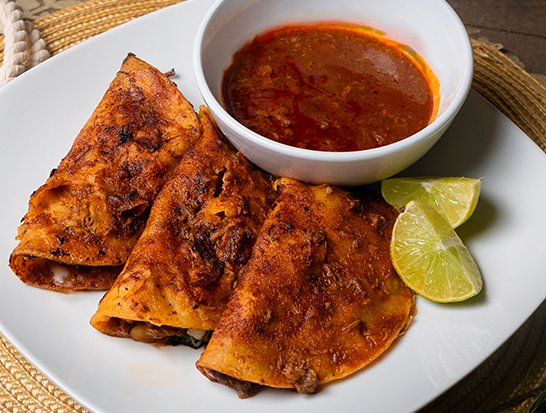 Delicious Quesabirrias - crispy tortillas filled with tender birria meat, served with rich consommé and fresh lime
